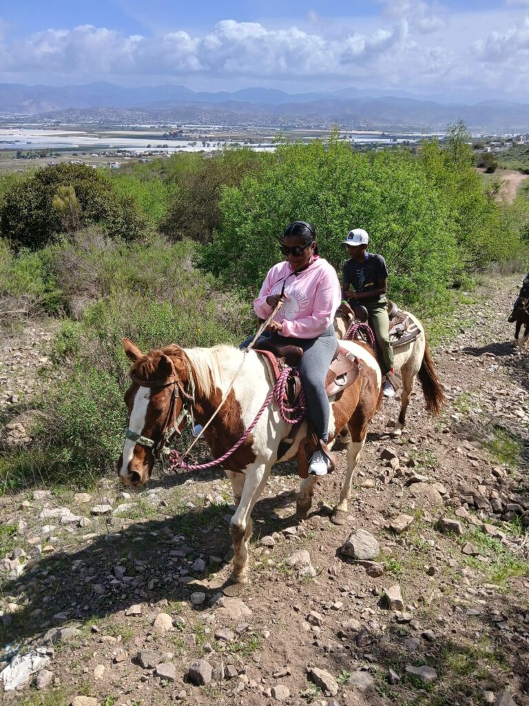 Two horseback riders facing the camera are on a desert trail with an overlook in the background.  Desert greenery lines the path.