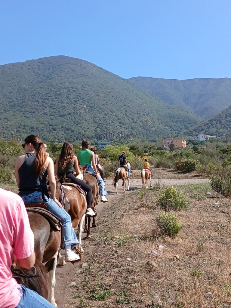 Several horseback riders follow a desert trail towards a green hill.