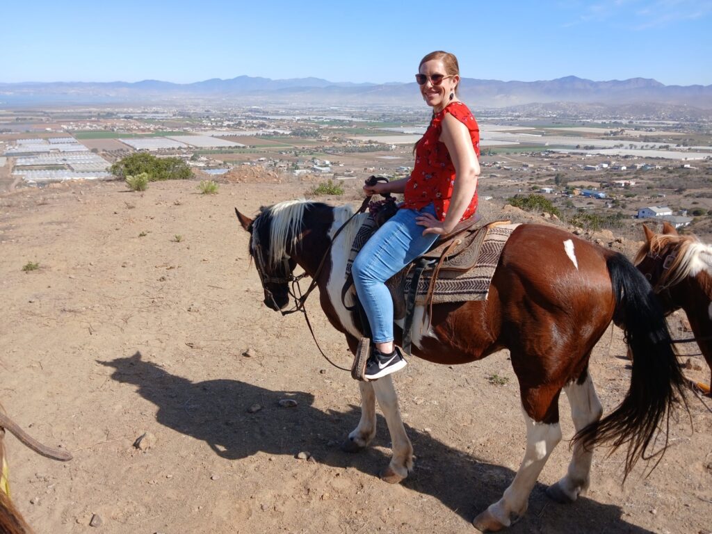 A woman sits on a horse, smiling and facing the camera.  Behind her is a panoramic view of Ensenada.