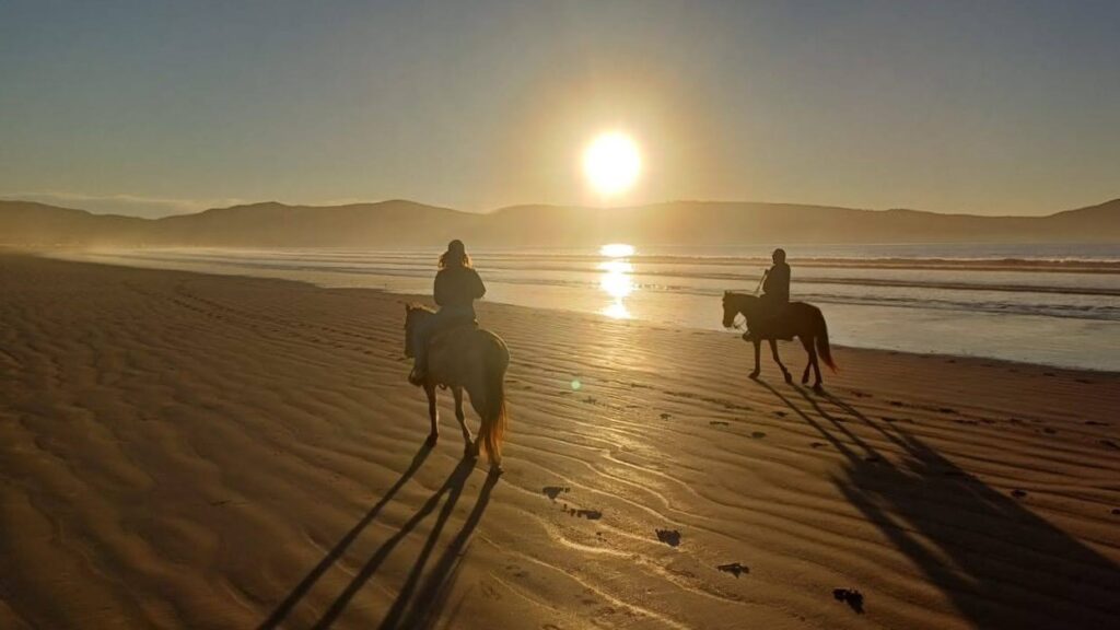 Two silhouetted horseback riders on a shallow beach shore, with mountains in the background and the sun setting over them.
