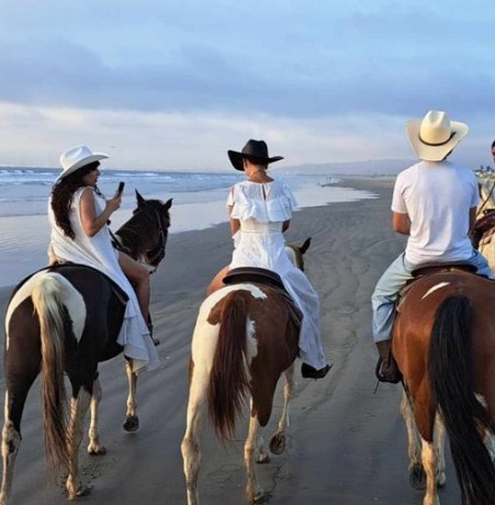 Three people wearing white and cowboy hats ride horses on a flat beach with mountains in the distance.